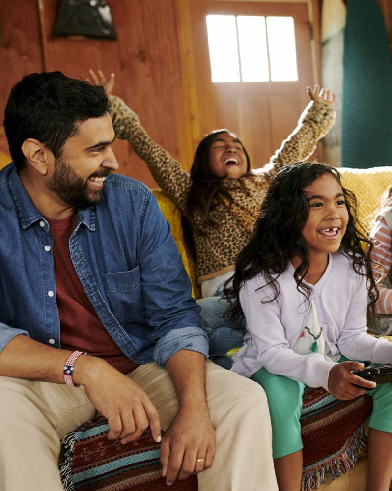 A smiling father watching his daughter play a game while another daughter behind them cheers.