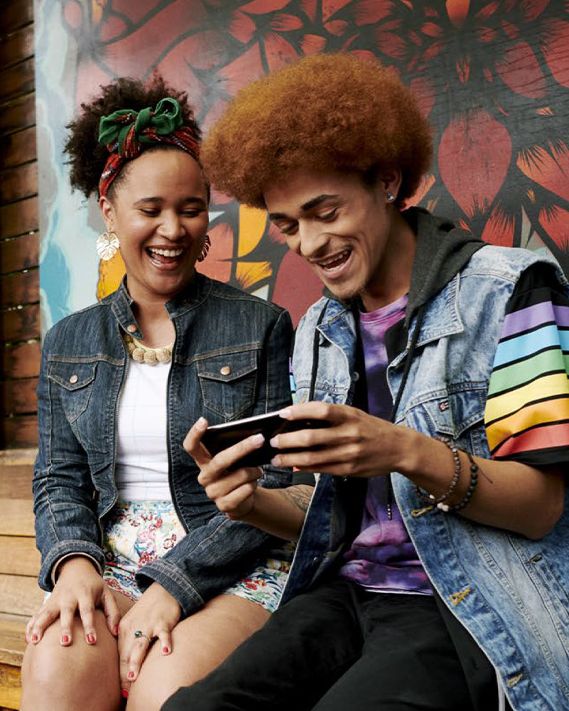 A man and woman are smiling as they watch him play a game on his phone in front of a large colorful mural.